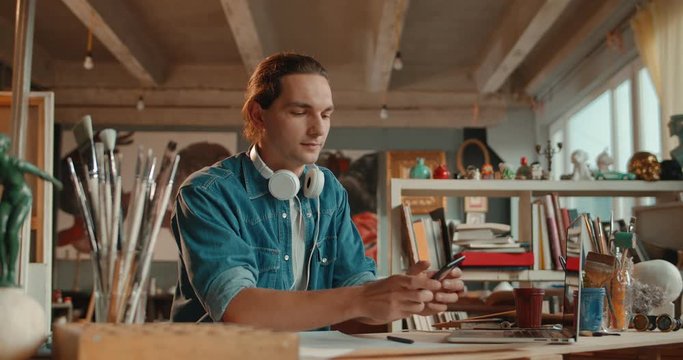 Caucasian young attractive guy artist typing and tapping on the smartphone while sitting at the desk in his cozy art studio.
