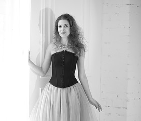 black and white portrait of a stylish model, girl in a tulle skirt and black corset is standing near the floor window. Beauty and fashion. Interior Design. Decor