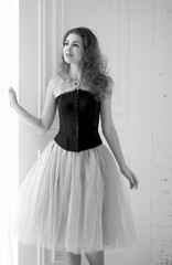 black and white portrait of a stylish model, girl  in a tulle skirt and black corset  looking out the window and  standing near the floor window. Beauty and fashion. Interior Design. Decor