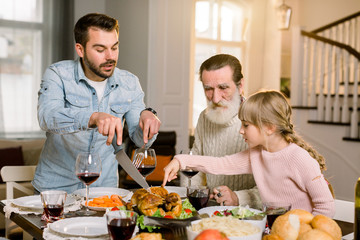 Image of handsome man father showing how to cut roasted turkey to his little cute daughter. Old grandfather, father and daughter having dinner together