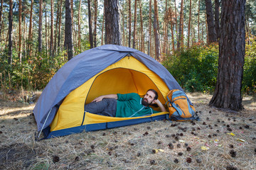 Handsome bearded young man listening to music in headphones while sitting in yellow tent in autumn forest. Hiking, travel, weekend trip concept. Healthy active lifestyle