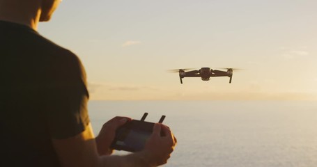 Young man piloting a drone at sunset flying out over the ocean, view of a man holding a drone controller and piloting his drone - Powered by Adobe