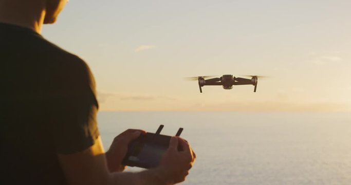 Young man piloting a drone at sunset flying out over the ocean, view of a man holding a drone controller and piloting his drone