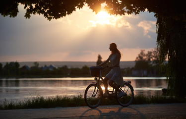 Silhouette of a young girl riding a bicycle along city lake at amazing summer sunset, rays of setting sun on evening sky, enjoying the nature, copy space
