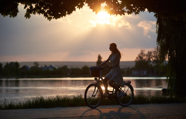 Fototapeta premium Silhouette of a young girl riding a bicycle along city lake at amazing summer sunset, rays of setting sun on evening sky, enjoying the nature, copy space