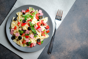 Traditional salad with pasta farfalle, ham, pepper and herbs on a dark plate on a dark background top view copy space