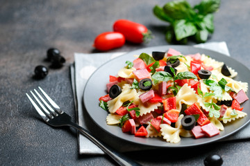 Traditional salad with pasta farfalle, ham, pepper and herbs on a dark plate on a dark background copy space