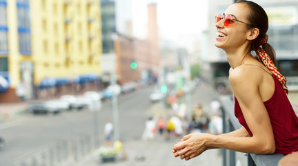 young smiling woman in trendy sunglasses and stylish red dress enjoying the city view from the balcony.
