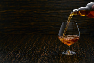 Pouring cognac from the bottle into the glass against wooden background.