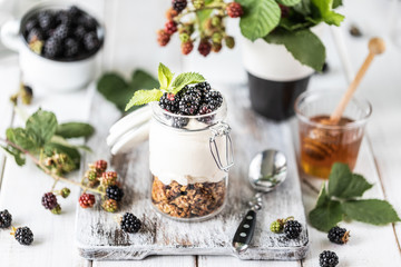 Handmade granola with white natural yogurt with blackberries in a glass transparent jar, flowers and leaves on a white wooden background.