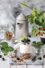 Handmade granola with white natural yogurt with blackberries in a glass transparent jar, flowers and leaves on a white wooden background.
