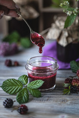 Glass jar with BlackBerry sauce close-up. Branch with berries and leaves in a wooden carved box on a dark wooden background.