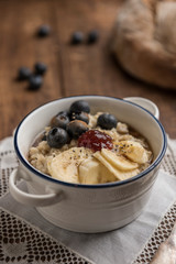 Porridge close-up with oat flakes, banana, blueberries, chia, cinnamon, maple syrup and strawberry jam on rustic wooden table. Country loaf in the background. Vertical. Selective focus
