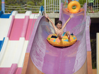 Summer fun in water park. Cute boy sliding down the slide on colorful inflatable floater.  His...