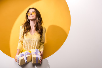 cheerful woman in sunglasses holding gifts on orange and white