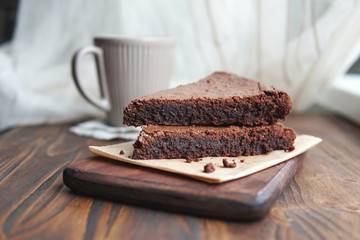 Freshly baked homemade chocolate cake - brownie on wooden board with cup of tea or coffee on rustic background. Selective focus.  Light from window. 