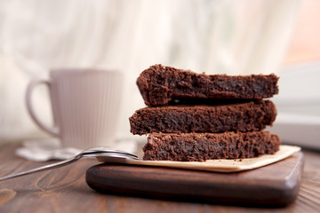 Freshly baked homemade chocolate cake - brownie on wooden board with cup of tea or coffee, spoons on rustic background. Selective focus.  Light from window. 