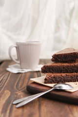 Freshly baked homemade chocolate cake - brownie on wooden board with cup of tea or coffee, spoons on rustic background. Selective focus.  Light from window. 