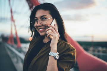 Cheerful Asian woman in trendy spectacles for vision protection smiling during friendly cellular conversation connected to 4g in roaming for discussing information, pretty young hipster girl - obrazy, fototapety, plakaty