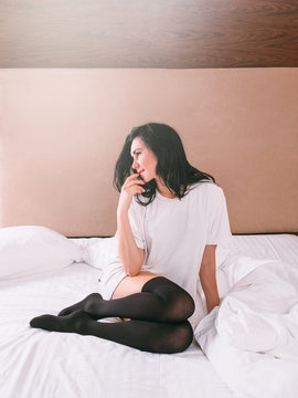 Dreamy, pensive brunette woman in white oversized boyfriend T shirt and black stockings sitting on bed in morning.