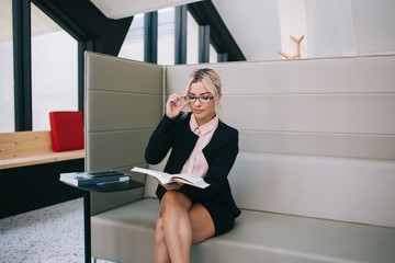 Concentrated female executive manager reading literature book with technology information, confident caucasian woman in stylish spectacles for vision correction interested on financial book