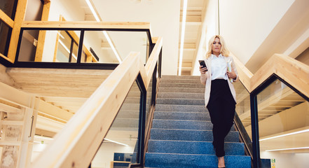 Full length portrait of serious confident female entrepreneur spending time in workspace going down on stairs for communicate with employees, attractive manager holding cellular phone in hand