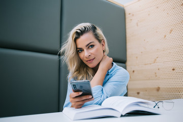 Young positive woman with blonde hair resting with literature best seller and modern cellular phone on publicity area, portrait of prosperous female student sitting with book and mobile indoors