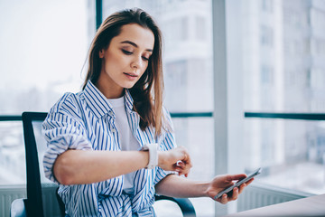 Young attractive female checking time of wrist watches while sitting in office interior holding...