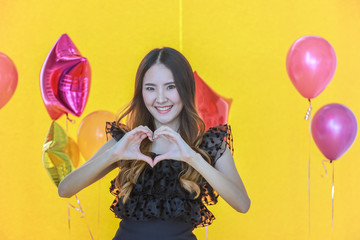 Portrait of smiling beautiful young asian woman use hand to show as heart shape with yellow wallpaper and colorfull balloon party.
