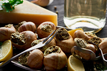 selective focus of delicious cooked escargots with lemon, parmesan, cutlery, parsley and white wine on black wooden table