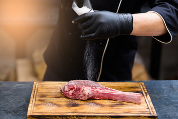Steakhouse kitchen. Cropped shot of chef cooking Cowboy steak, salting raw beef bone in meat on...