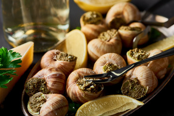 selective focus of delicious cooked escargots with lemon, parmesan, cutlery, parsley and white wine on black wooden table