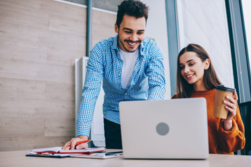 Cheerful male and female partners searching interesting information for business startup project during cooperation in office sitting at desktop with takeaway coffee, concept of modern technology