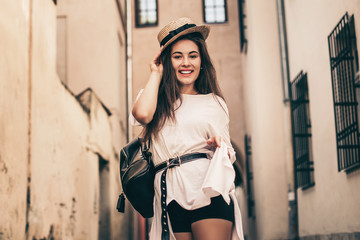 Young stylish woman walking on the old town street, travel with backpack, straw hat, wearing trendy outfit.