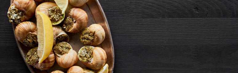 top view of delicious cooked escargots with lemon on black wooden table, panoramic shot
