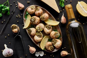 top view of delicious cooked escargots with lemon slices on black wooden table with spices, Parmesan and white wine