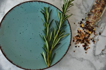 sprigs of rosemary in a blue round plate and sprinkled seasonings with pepper and salt on a white marble background