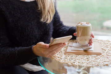 Woman drink Hot latte or Cappuccino coffee in a cafe near the window using mobile phone. Relax, coffee break, breakfast, morning, coworking, freelance.
