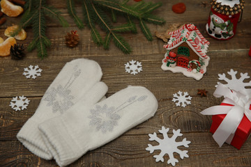 Obraz na płótnie Canvas white mittens with a snowflake made of rhinestones on a background of Christmas toys, a gingerbread house and fir branches on a wooden table. The concept of Christmas and New Year.