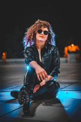 Fototapeta na wymiar A young pretty girl with curly hair sitting on the floor, wearing sunglasses in a night shooting, looking straight ahead