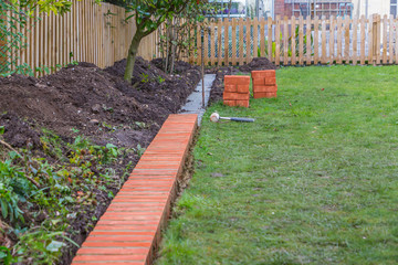 A string line keeping installation of a brick edging to a lawn straight. The cement footings can be seen where new bricks are stacked waiting for laying.