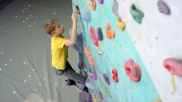 Tracking shot of 13-year-old blond Caucasian boy in yellow t-shirt, pants and shoes climbing up indoor vertical wall with manmade holds towards top, and padded crash mats on floor below
