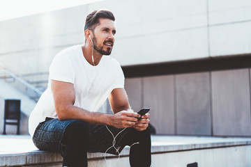 Handsome male runner dressed in stylish apparel looking away while listening audio advices for training connected to 4g internet on cellphone, man in sportswear sitting on urban setting with phone