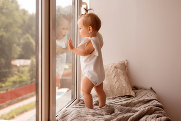 baby girl 11 months stands on the windowsill and looks out the window. warmth and comfort in the house. sound insulation and plastic Windows in the house