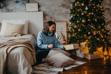 Young woman sits next to Christmas tree and holds box with gift.