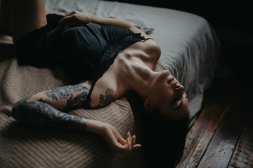 Young woman in black lingerie with tattoo on her arm lies on bed.