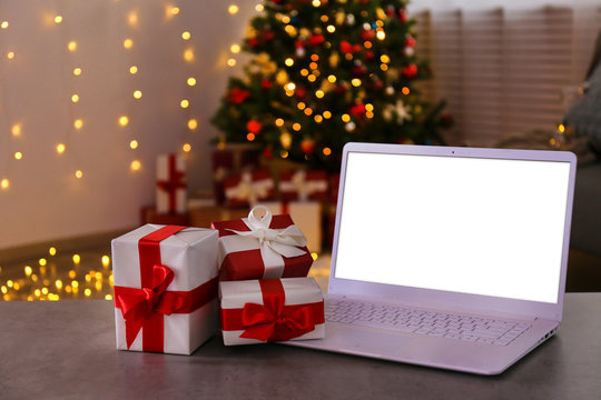 Blank screen laptop. Traditional christmas pine tree with holiday decorations & stack of presents. Bokeh lights glowing in the evening. Close up, copy space, background, interior.