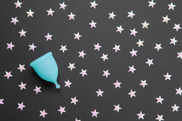 Turquoise menstrual cup on black background with numerous stars. Concept possibility of use cup at night, zero waste, savings, minimalism. Feminine hygiene product, flat lay, copy space. Horizontal
