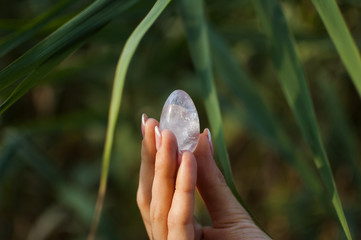 Female hand with french manicure holding transparent violet amethyst yoni egg for vumfit, imbuilding or meditation. Crystal quartz egg in hands on green background outdoors