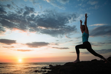 Yoga woman doing exercises on the ocean during amazing sunset.